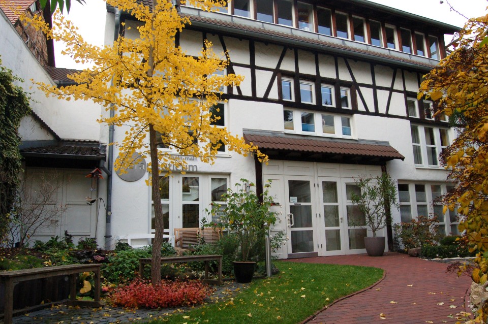 Ein weißes Fachwerkhaus mit dunkelbraunem Holzrahmen und großen Fenstern, umgeben von herbstlich gefärbtem Laub und einer gepflegten Rasenfläche mit rotem Backsteinweg.