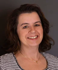 Martina Ecker, Portraitfoto einer lächelnden Frau mit schulterlangen braunen Wellen und einem graumeliertem Strickpullover über einem dunklen Hintergrund.