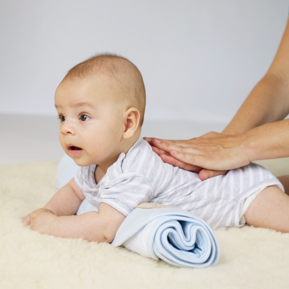 Ein Baby in hellblauem Hemd liegt auf einer cremefarbenen Oberfläche, während eine Hand es am Rücken stützt. Das Kind blickt überrascht zur Seite und trägt eine blaugestreifte Windel.