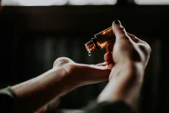 Eine Person gießt ein braunes Öl oder eine Flüssigkeit aus einer Flasche in die offene Hand einer anderen Person. Die Aufnahme zeigt eine Nahansicht mit natürlicher Beleuchtung vor dunklem Hintergrund.