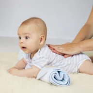 Ein Säugling in hellblauem gestreiftem Hemd liegt auf einer hellen Decke und wird von einer erwachsenen Hand sanft am Rücken berührt. Das Baby blickt aufmerksam zur Seite, während eine blaue Windel neben ihm liegt.