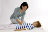 Eine Frau führt eine Shiatsu-Massage an einem entspannten Kind durch, das auf dem Rücken liegt. Das Kind trägt ein blau-weiß gestreiftes Shirt, während die Frau sanft Druck auf seinen Bauch ausübt. Die Szene zeigt eine therapeutische Behandlung in ruhiger, heller Umgebung.