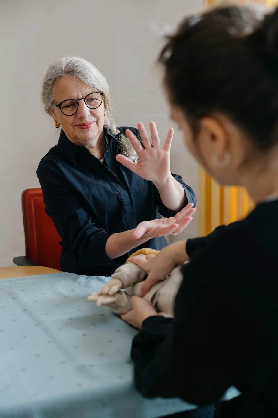 Eine ältere Frau mit Brille gestikuliert lächelnd, während eine Person im Vordergrund eine Handpuppe hält.