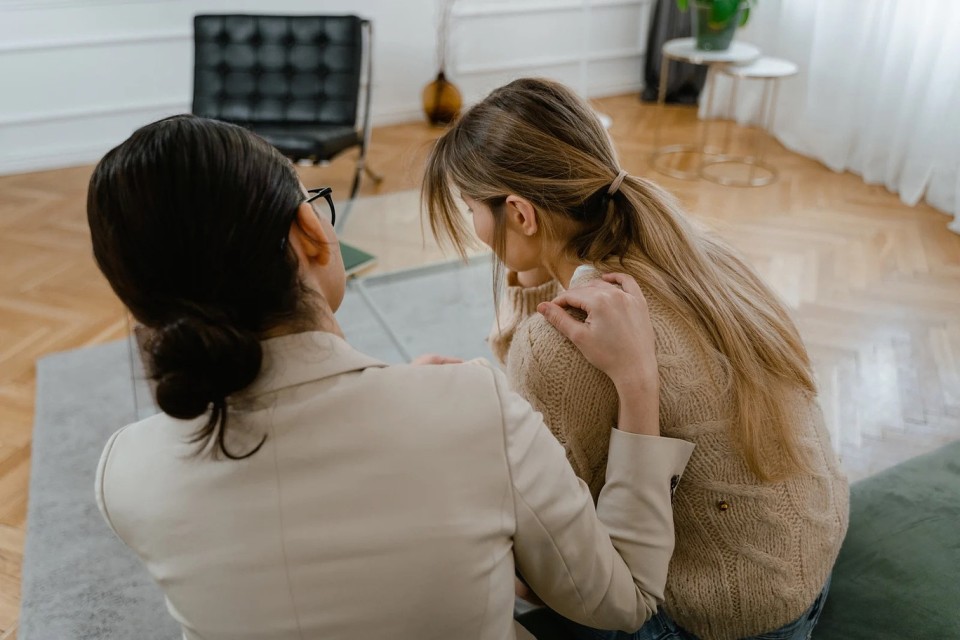 Zwei Frauen sitzen sich auf dem Boden eines hellen Wohnzimmers gegenüber und führen ein intensives Gespräch. Die Frau auf der rechten Seite wirkt emotional bewegt und hält sich die Hand vor das Gesicht.