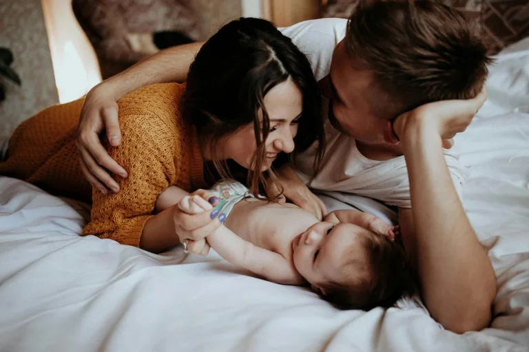 Ein junges Paar liegt auf weißer Bettwäsche und umarmt zärtlich sein neugeborenes Baby. Die Mutter in orangegelbem Shirt und der Vater in hellem Shirt schauen liebevoll auf ihr Kind herab.