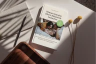 Ein Buch mit dem Titel "Warum manche Eltern Sicherheit brauchen" liegt auf einem weißen Untergrund neben getrockneten Blüten und schwarzen Stiften. Das Cover zeigt ein Foto von zwei Kindern. Ein grüner Kreis mit Text ist auf dem Cover sichtbar.