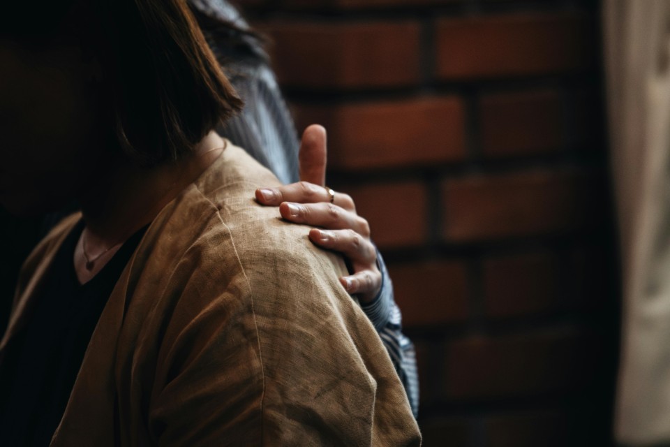 Eine schwangere Frau berührt sanft ihren Bauch mit beiden Händen. Sie trägt ein helles, langärmeliges Oberteil und steht vor einer Backsteinmauer. Das warme Licht unterstreicht die zärtliche Geste.