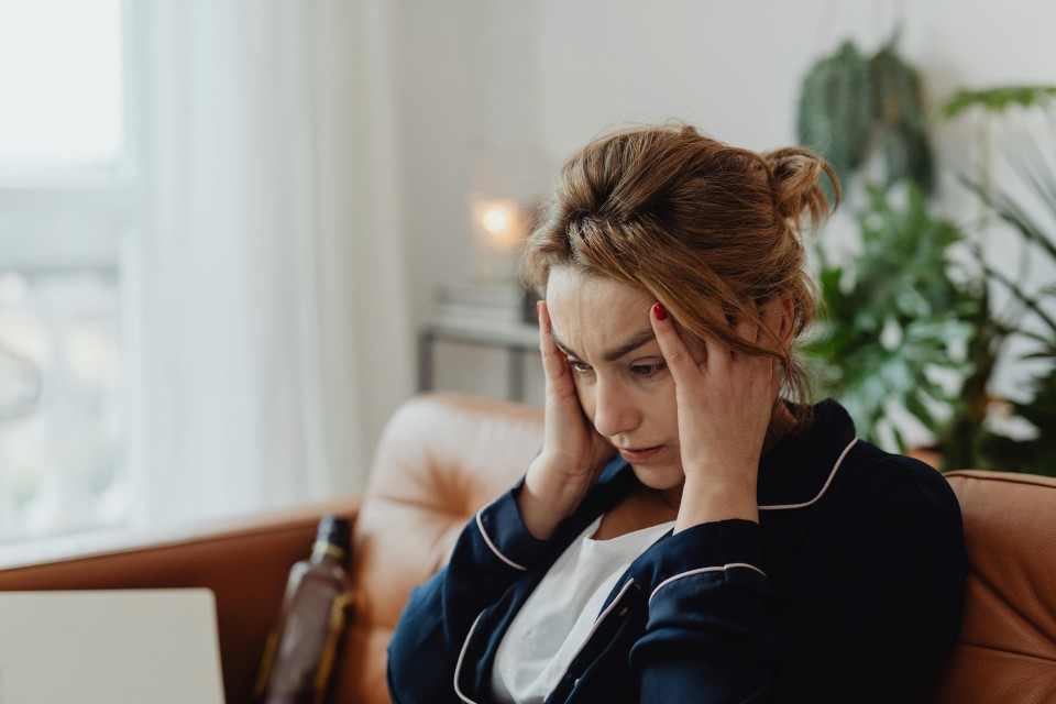 Eine Frau mit hochgestecktem Haar sitzt auf einem Sofa und stützt nachdenklich ihr Gesicht in die Hand. Sie trägt einen dunklen Blazer über einem hellen Shirt und schaut gedankenverloren nach unten. Im Hintergrund sind ein helles Fenster und eine Grünpflanze sichtbar.