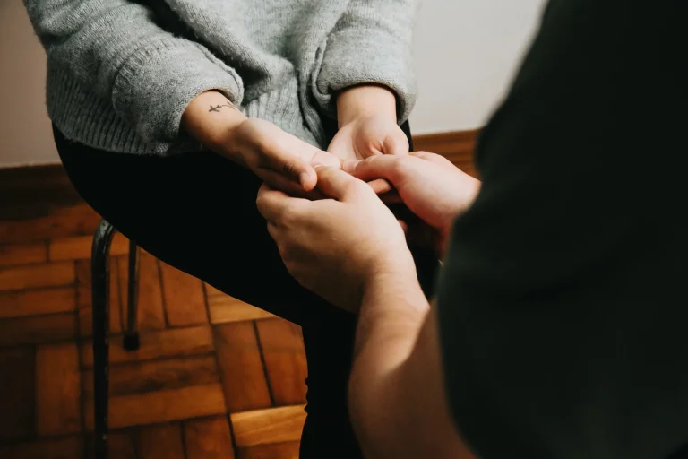 Zwei Personen halten sich an den Händen in einem Zeichen von Unterstützung und Verbundenheit. Die Hände ruhen auf einer Holzoberfläche, während eine Person einen grauen Pullover trägt. Die Geste symbolisiert Zusammenhalt und gegenseitige Hilfe.