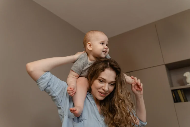 Eine Frau mit langen braunen Haaren trägt ein kleines Kind auf ihren Schultern, das sie mit beiden Armen hält. Beide schauen nach oben und lächeln in einem hellen, modernen Wohnraum.