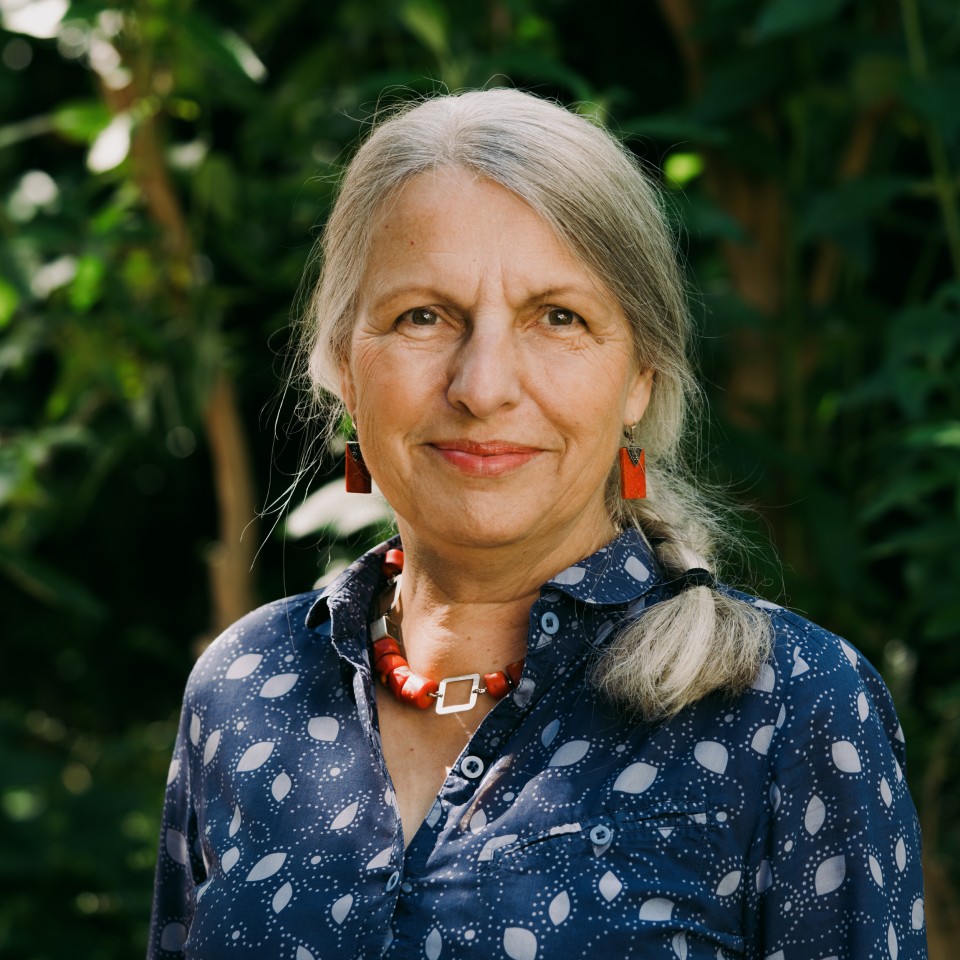 Eine Frau mit grauem, schulterlangem Haar steht vor grünem Laub. Sie trägt ein dunkelblau-weißes Hemd, eine rote Halskette und rote Ohrringe und blickt direkt in die Kamera.