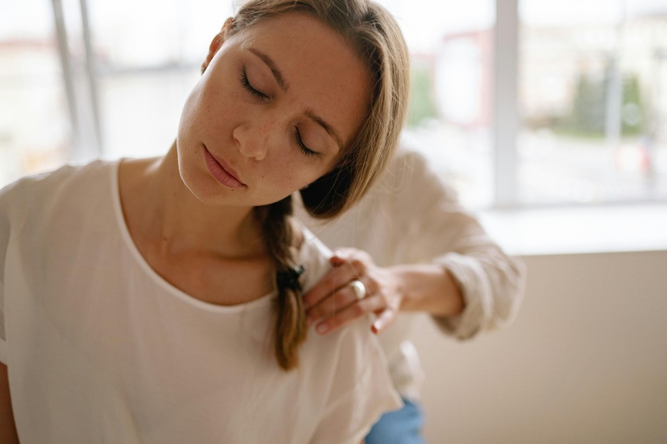 Eine lächelnde Frau mit blonden Haaren hält zärtlich ein kleines weißes Kätzchen in ihren Armen. Sie trägt ein helles Langarmshirt und sitzt in einem hellen Raum mit Fenstern im Hintergrund. Ihr Blick ist liebevoll auf das Tier gerichtet.