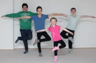 Vier Kinder in verschiedenfarbigen Shirts führen eine Balanceübung auf einem Bein durch, während sie ihre Arme ausgestreckt halten. Sie stehen vor einer weißen Wand und demonstrieren Körperkontrolle und Koordination.