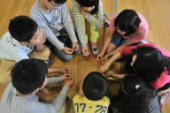 Eine Gruppe von Kindern sitzt im Kreis auf einem Holzboden und legt ihre Hände zusammen in der Mitte. Die Kinder tragen verschiedenfarbige Kleidung und schauen nach unten auf ihre verbundenen Hände.