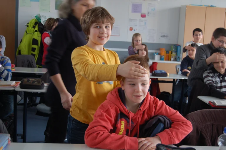 Ein Kind in gelbem Pullover legt seine Hand auf den Kopf eines anderen Kindes in rotem Pullover, das lächelt. Sie sitzen in einem Klassenzimmer mit anderen Kindern im Hintergrund.
