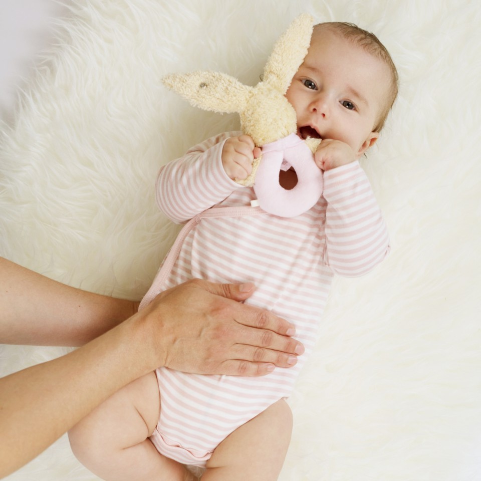 Ein Baby in einem rosa-weiß gestreiften Strampler liegt auf einer weißen, flauschigen Oberfläche und hält ein cremefarbenes Stofftier in der Hand, während es von erwachsenen Händen gehalten wird.