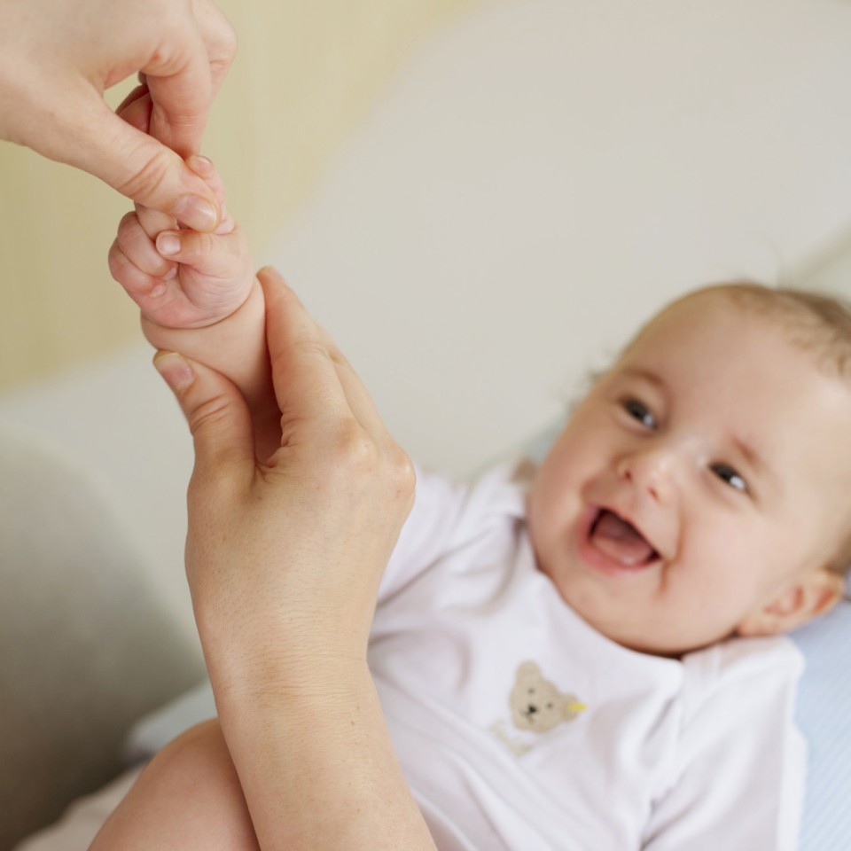 Ein lächelndes Baby in weißem Shirt hält die Hand eines Erwachsenen und greift mit seinen Fingern zu. Die beiden Hände zeigen eine liebevolle Verbindung zwischen Elternteil oder Betreuer und Kind.