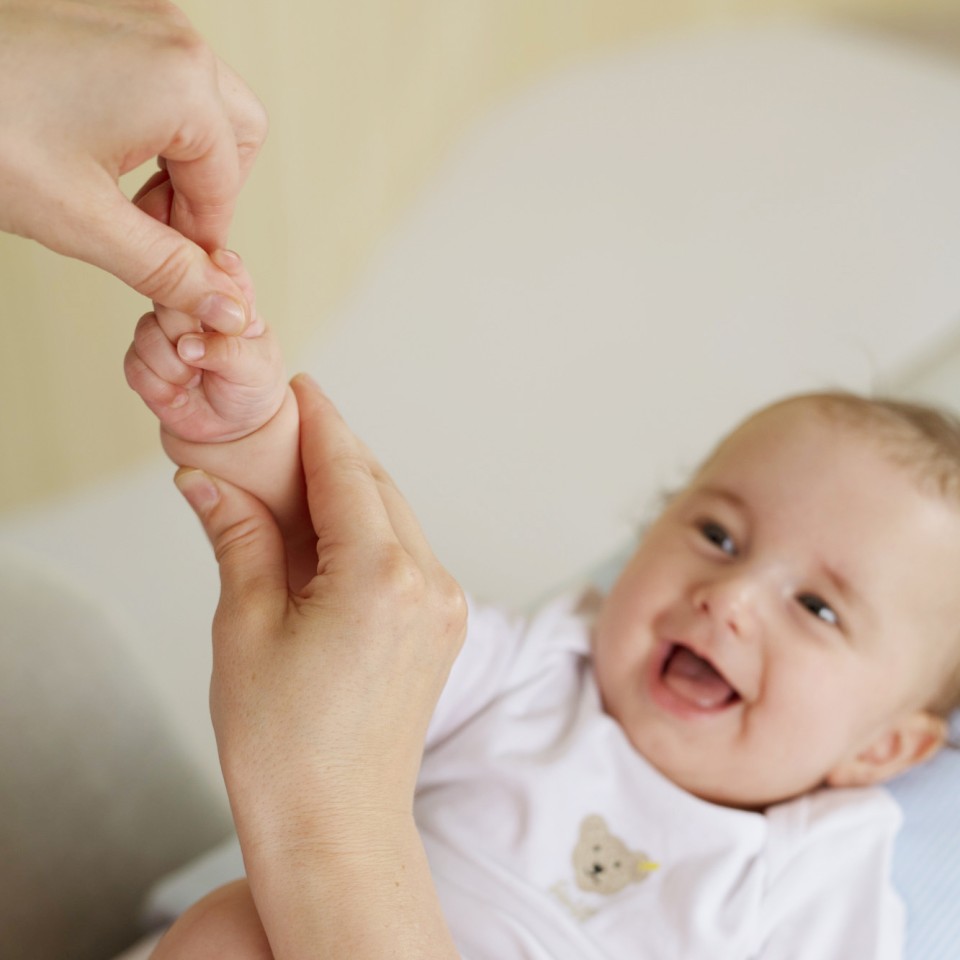 Ein lächelndes Baby in weißem Oberteil hält die Finger eines Erwachsenen und schaut zur Seite. Die Szene zeigt eine liebevolle Interaktion zwischen Erwachsenem und Kleinkind.