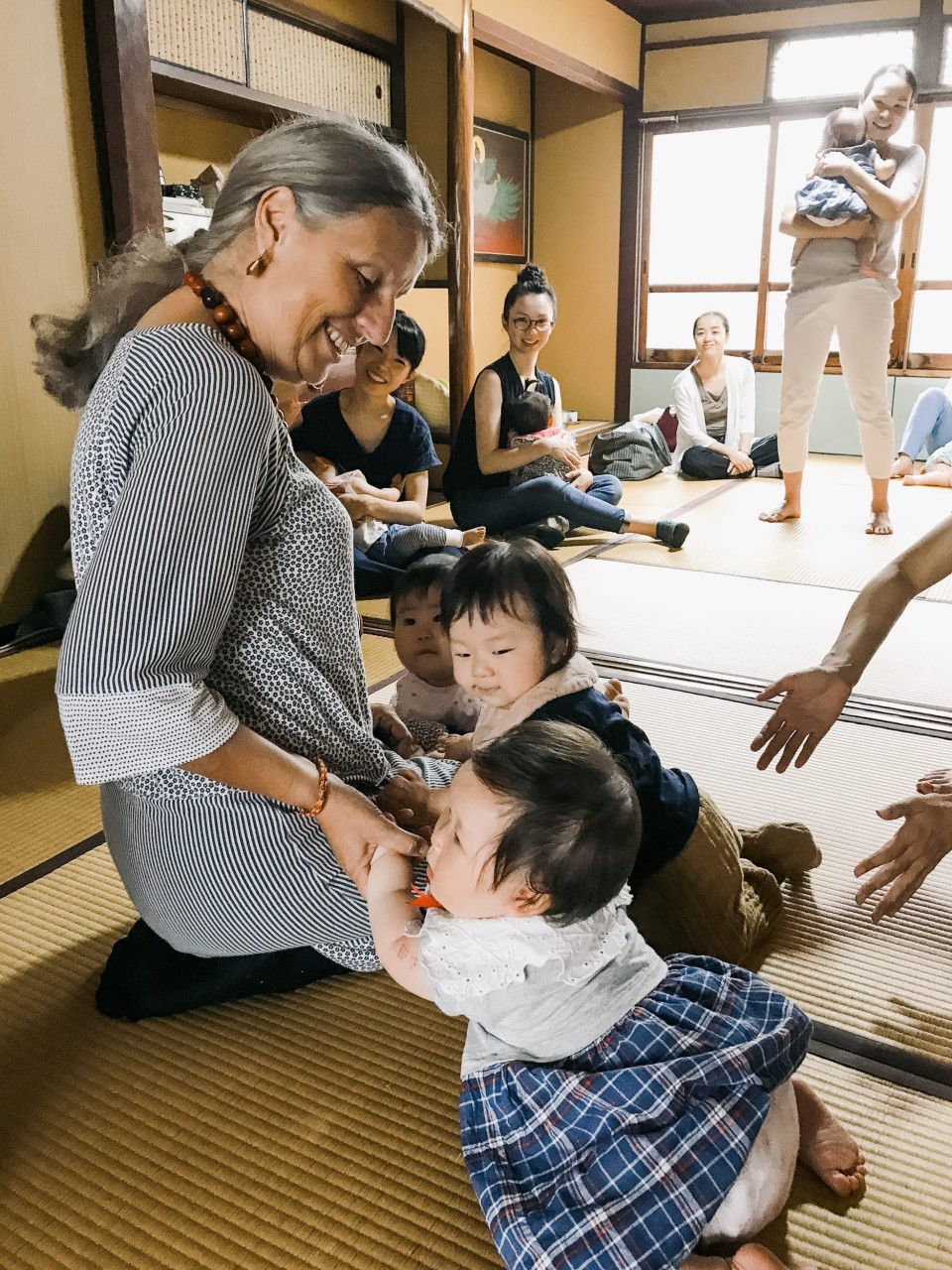 Eine Frau kniet neben zwei kleinen Kindern und demonstriert eine Shiatsu-Massage-Technik während eines Workshops in einem traditionellen japanischen Raum. Im Hintergrund sitzen weitere Teilnehmer und beobachten die praktische Anwendung.