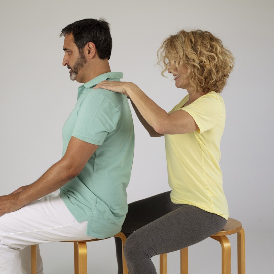 Ein Mann sitzt auf einem Hocker und wird von einer Frau von hinten massiert oder behandelt. Beide tragen helle Freizeitkleidung und sitzen sich gegenüber auf Hockern vor weißem Hintergrund.