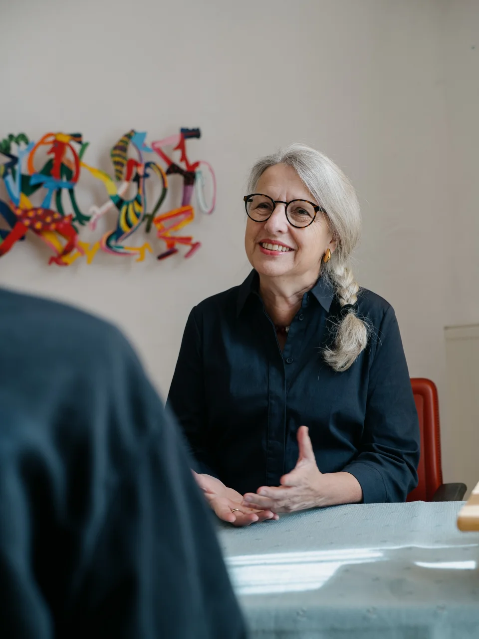 Frau mit grauem Zopf und Brille lächelt und spricht in einem Gespräch an einem Tisch