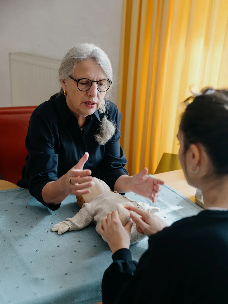 Ältere Frau mit grauen Zöpfen und Brille zeigt einer jungen Frau an einer Babypuppe, wie sie mit den Händen arbeiten soll.