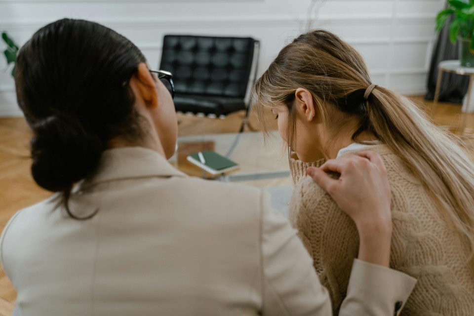 Zwei Frauen sitzen sich in einem hellen Büro gegenüber und führen ein Gespräch. Im Hintergrund sind ein Laptop, ein schwarzer Stuhl und eine Pflanze zu sehen.