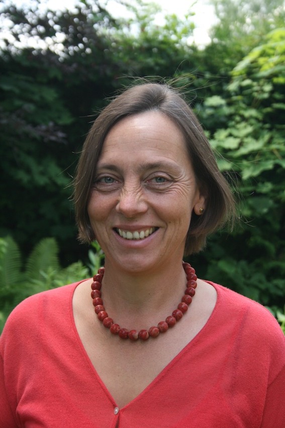 Eine Frau mit schulterlangen braunen Haaren lächelt in die Kamera. Sie trägt ein korallenfarbenes Shirt und eine rote Perlenkette. Im Hintergrund ist grünes Laub zu sehen.