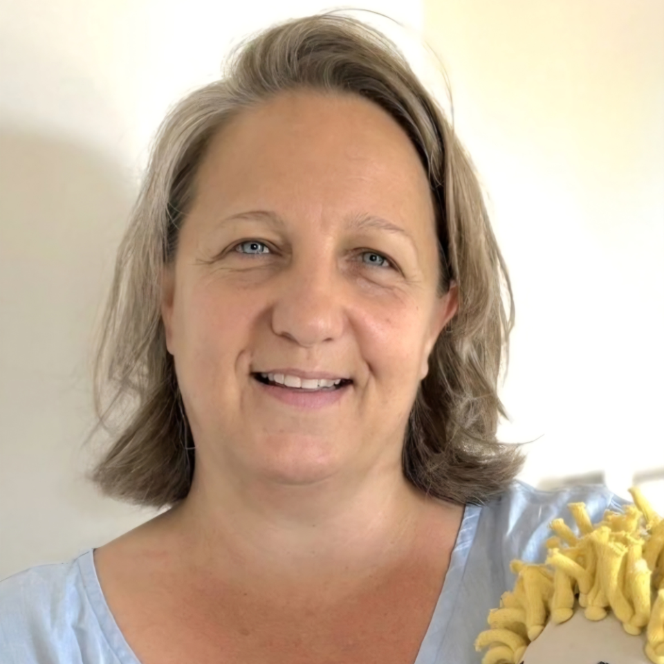 Eine Frau mit schulterlangem grauem Haar lächelt in die Kamera. Sie trägt ein hellblaues Shirt und hält ein gelbes Objekt in der Hand. Der Hintergrund ist hell und neutral.