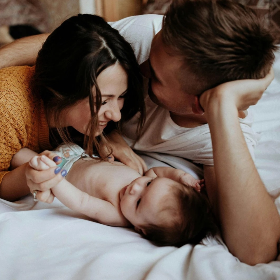 Zwei Eltern liegen auf einem Bett und spielen liebevoll mit ihrem kleinen Baby, das zwischen ihnen liegt. Sie schauen sich gegenseitig an und interagieren zärtlich mit dem Neugeborenen in einer warmen, familiären Atmosphäre.