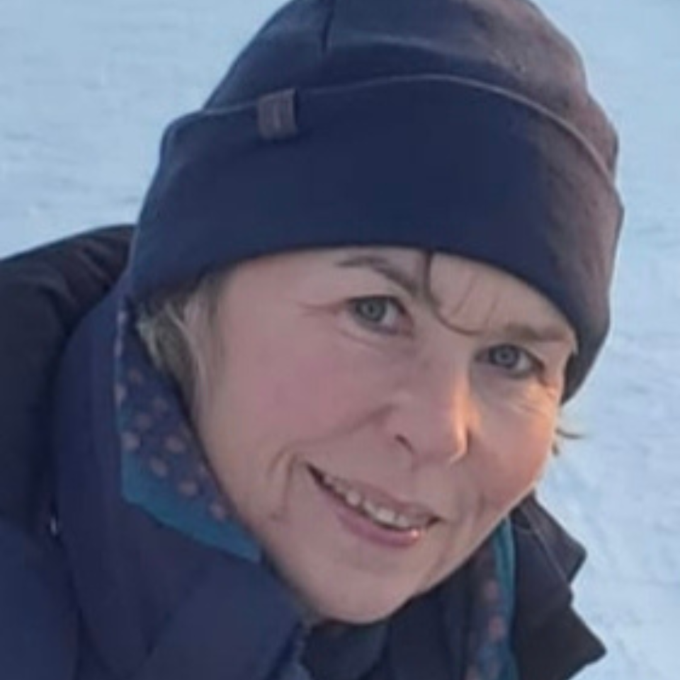 Eine Frau mit dunkler Mütze und schwarzer Jacke lächelt in die Kamera. Im Hintergrund ist eine schneebedeckte Landschaft zu sehen. Sie trägt einen freundlichen, offenen Gesichtsausdruck.