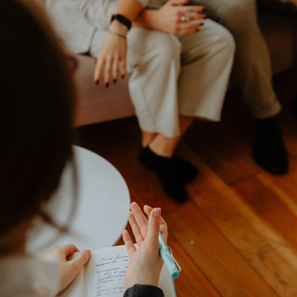 Mehrere Personen sitzen zusammen auf einem Holzboden und halten gemeinsam ein Blatt Papier, auf dem handschriftliche Notizen zu sehen sind. Die Hände der Teilnehmer sind sichtbar, während sie das Dokument betrachten und besprechen.