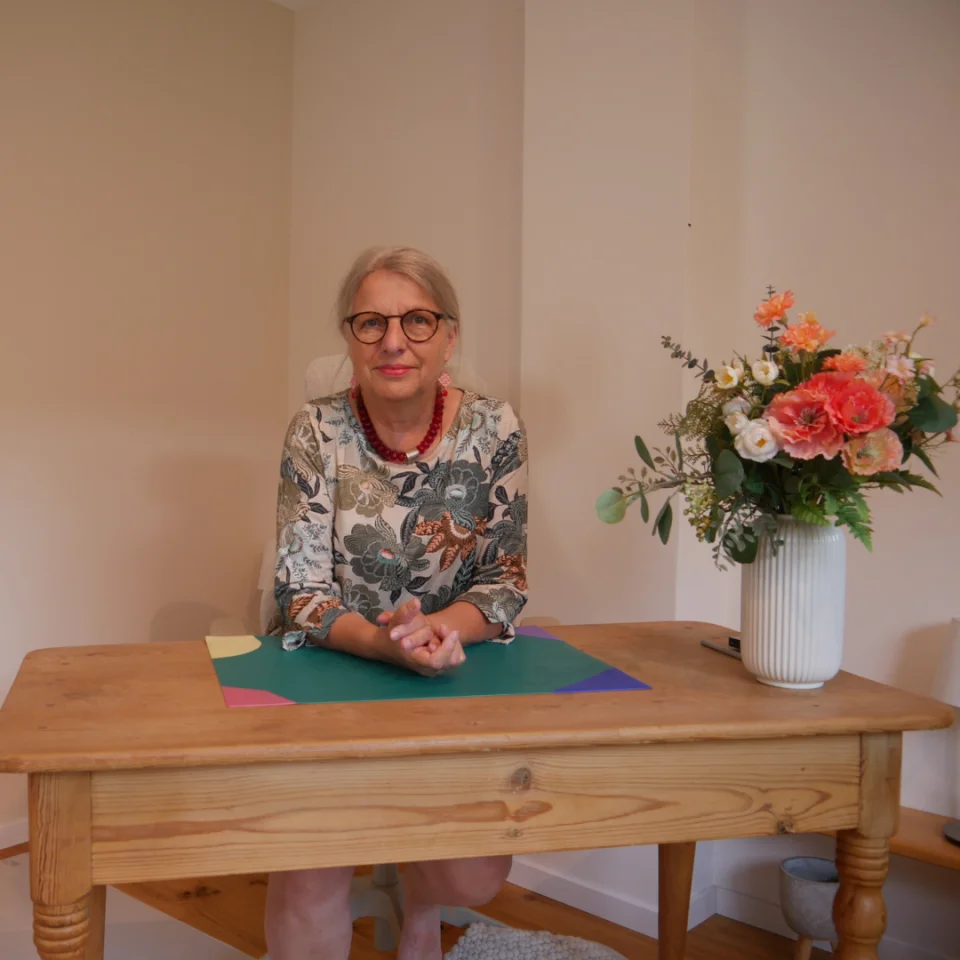 Eine Frau mit Brille sitzt an einem Holztisch und legt ihre Hände auf bunte Papierblätter. Neben ihr steht eine weiße Vase mit einem bunten Blumenstrauß auf dem Tisch.