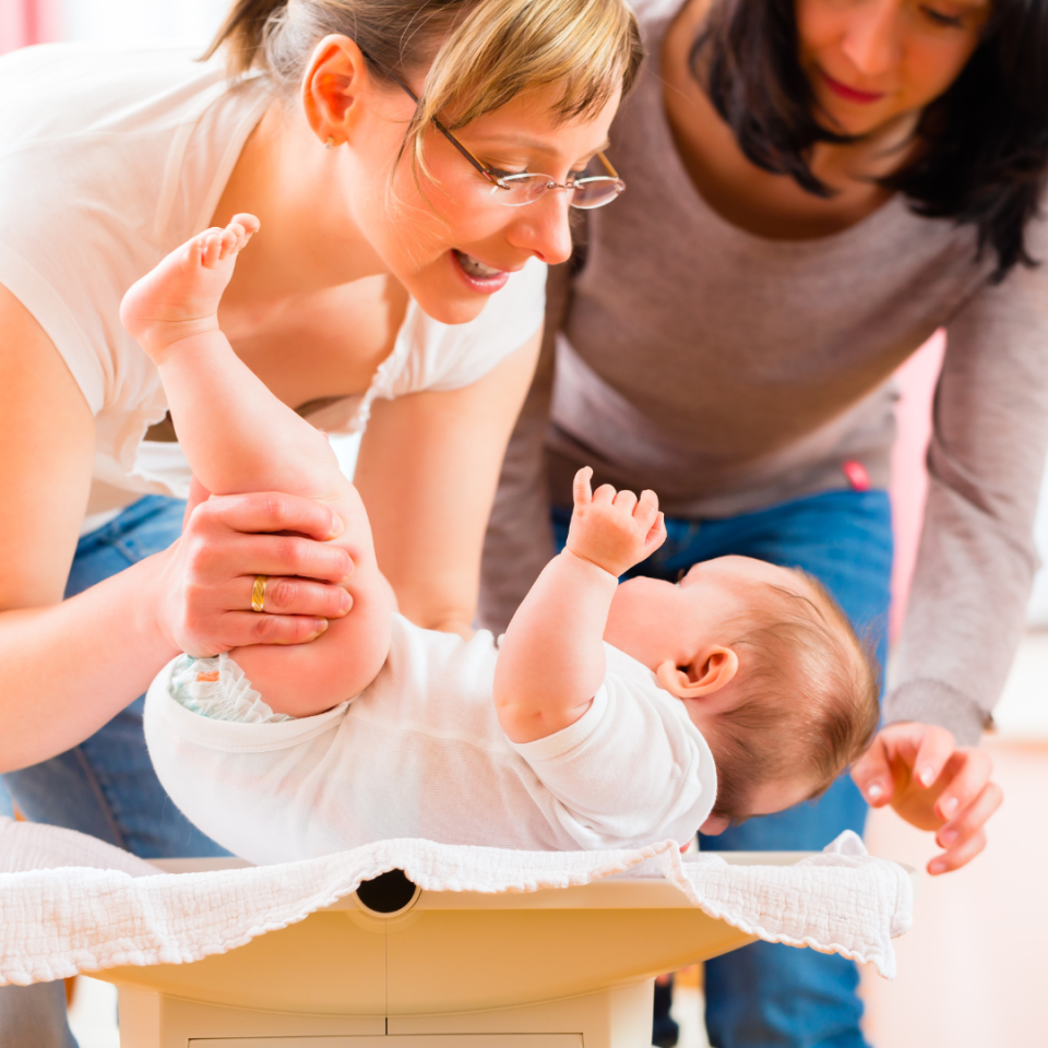 Zwei Frauen und ein Baby bei einer Bewegungsübung auf einer Gymnastikmatte. Eine Frau in Brille und weißem Shirt hält das Baby, während die andere Frau daneben sitzt und beobachtet. Das Baby liegt auf dem Rücken und macht aktive Bewegungen mit Armen und Beinen.