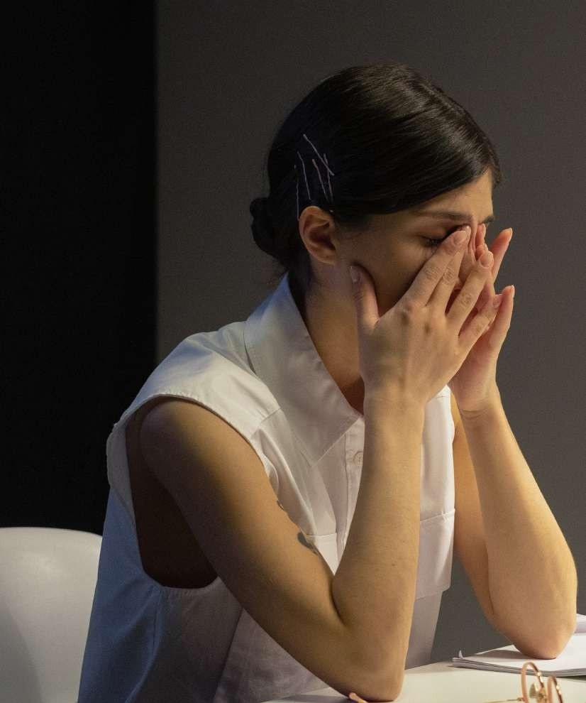 Eine Frau sitzt an einem Tisch und stützt ihr Gesicht mit beiden Händen, während sie nachdenklich nach unten blickt. Sie trägt ein weißes ärmeloses Oberteil und wirkt konzentriert oder besorgt. Die Beleuchtung ist gedimmt und schafft eine ernsthafte Atmosphäre.