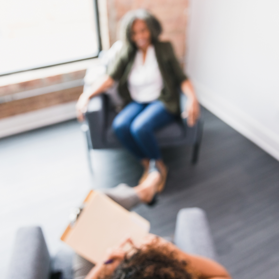 Eine Frau sitzt auf einem grauen Sessel in einem Beratungszimmer und blickt auf eine Person, deren Schulter im Vordergrund zu sehen ist. Im Hintergrund sind ein Whiteboard und helle Wände erkennbar.