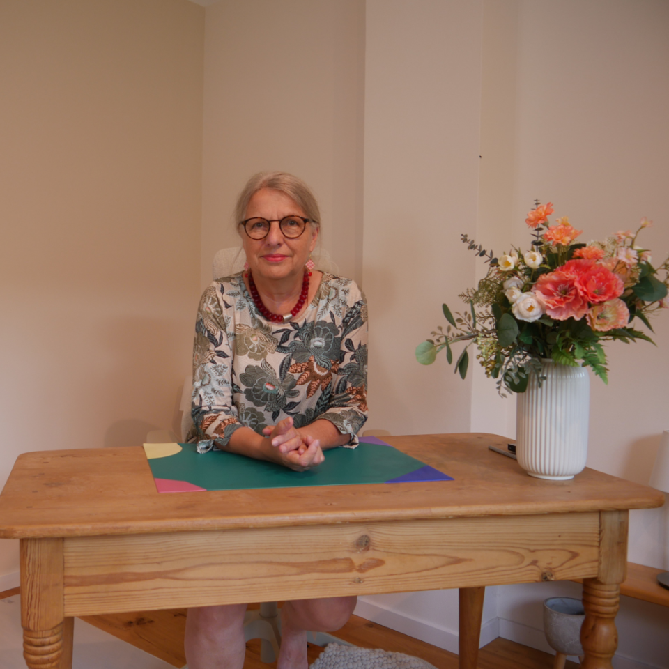 Eine ältere Frau mit Brille und grauem Haar sitzt an einem Holztisch und legt ihre Hände auf farbige Papiere. Neben ihr steht eine weiße Vase mit roten und orangefarbenen Blumen. Im Hintergrund sind cremefarbene Wände sichtbar.