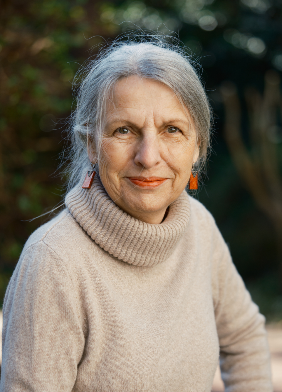 Eine Frau mit grauem Haar und orangefarbenen Ohrrings trägt einen beigen Rollkragenpullover und schaut direkt in die Kamera. Sie hat ein ruhiges, direktes Gesicht mit leichtem Lächeln vor unscharfem grünem Hintergrund.