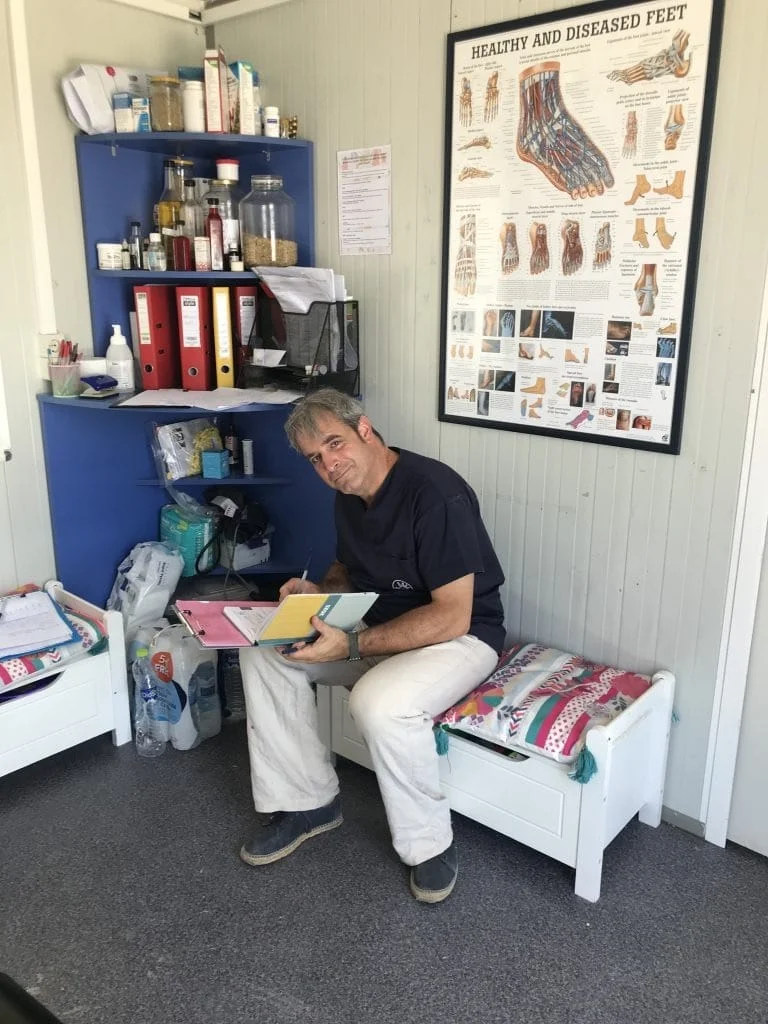 Ein Mann sitzt in einer Klinik oder Behandlungsraum und hält medizinische Materialien in den Händen. Hinter ihm steht ein blaues Regal mit medizinischen Vorräten und an der Wand hängt ein anatomisches Poster über Fußerkrankungen. Der Raum ist funktional eingerichtet als medizinische Einrichtung.
