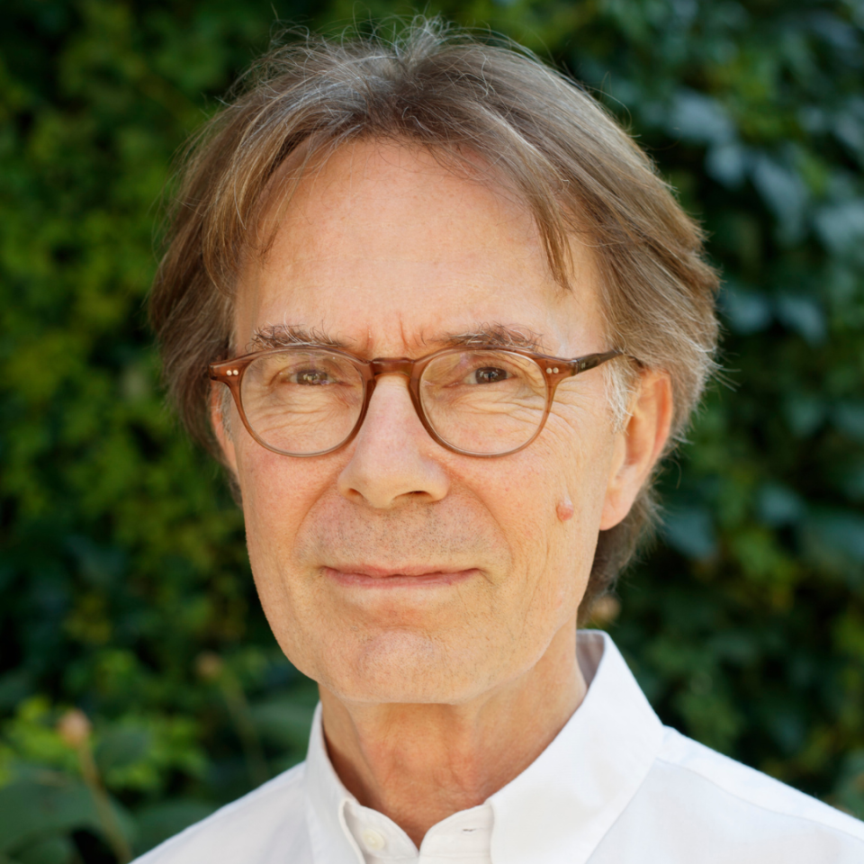 Ein Mann mittleren Alters mit grauem Haar und Brille trägt ein weißes Hemd vor einem grünen Hintergrund aus Blattwerk.