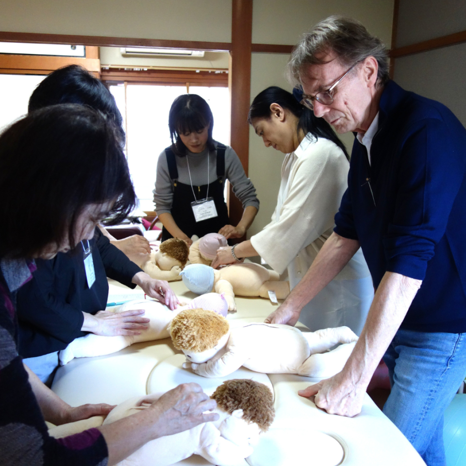 Ein Mann und vier Frauen untersuchen während eines Workshops verschiedene getrocknete Kräuter und Materialien auf weißen Tellern. Die Teilnehmer tragen Namensausweise und konzentrieren sich auf die praktische Demonstration von Shonishin-Techniken und Materialien.