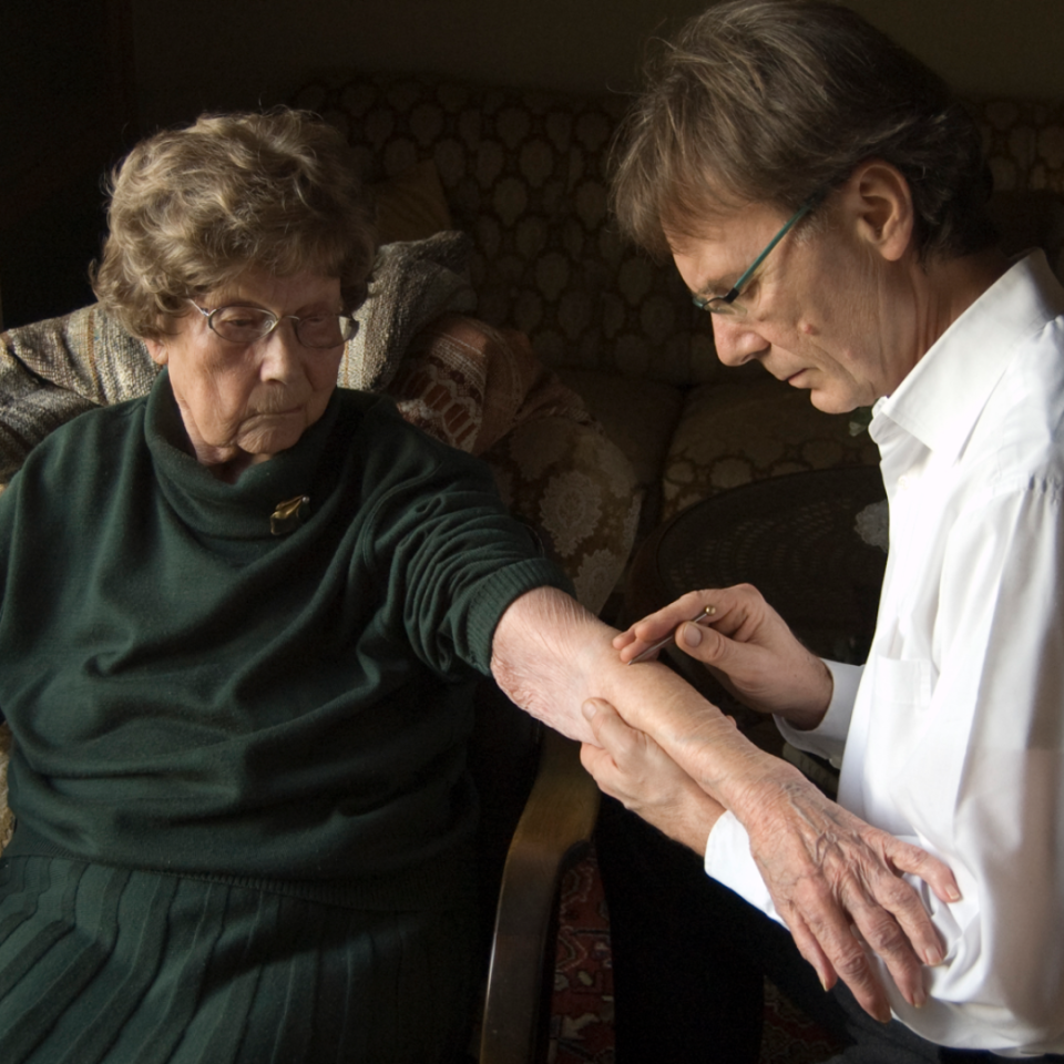 Eine Therapeutin in weißem Hemd untersucht die Hand einer älteren Frau in grünem Pullover. Beide tragen Brillen und sitzen sich gegenüber. Die Szene zeigt eine klinische Untersuchung oder Behandlung im Rahmen der Shonishin-Ausbildung.